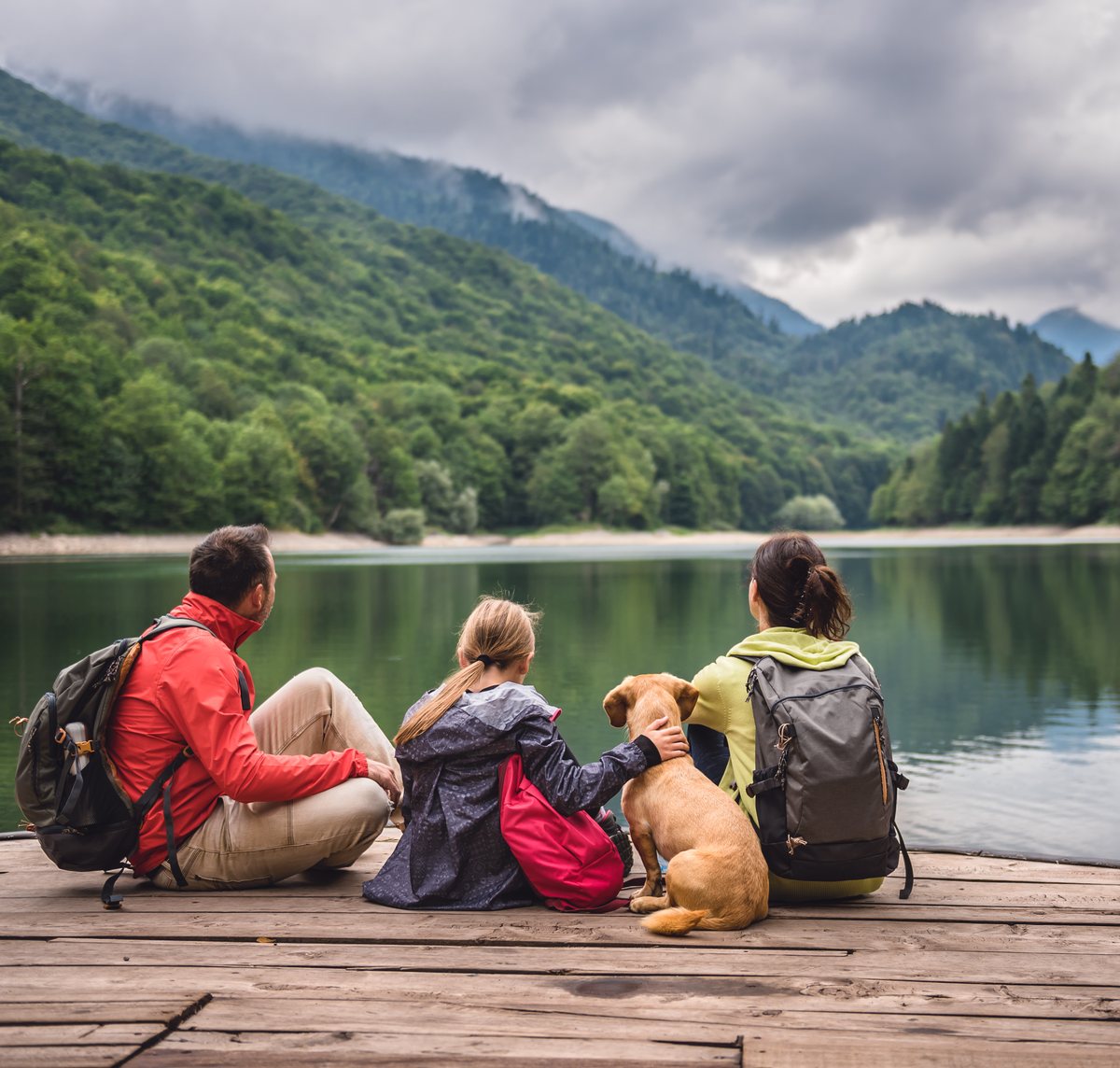 Family with a dog sitting together on a dock, looking out over a calm mountain lake
